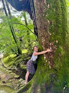 Natalia Nobes outdoors in a mossy forest, resting a hand against the trunk of an old tree